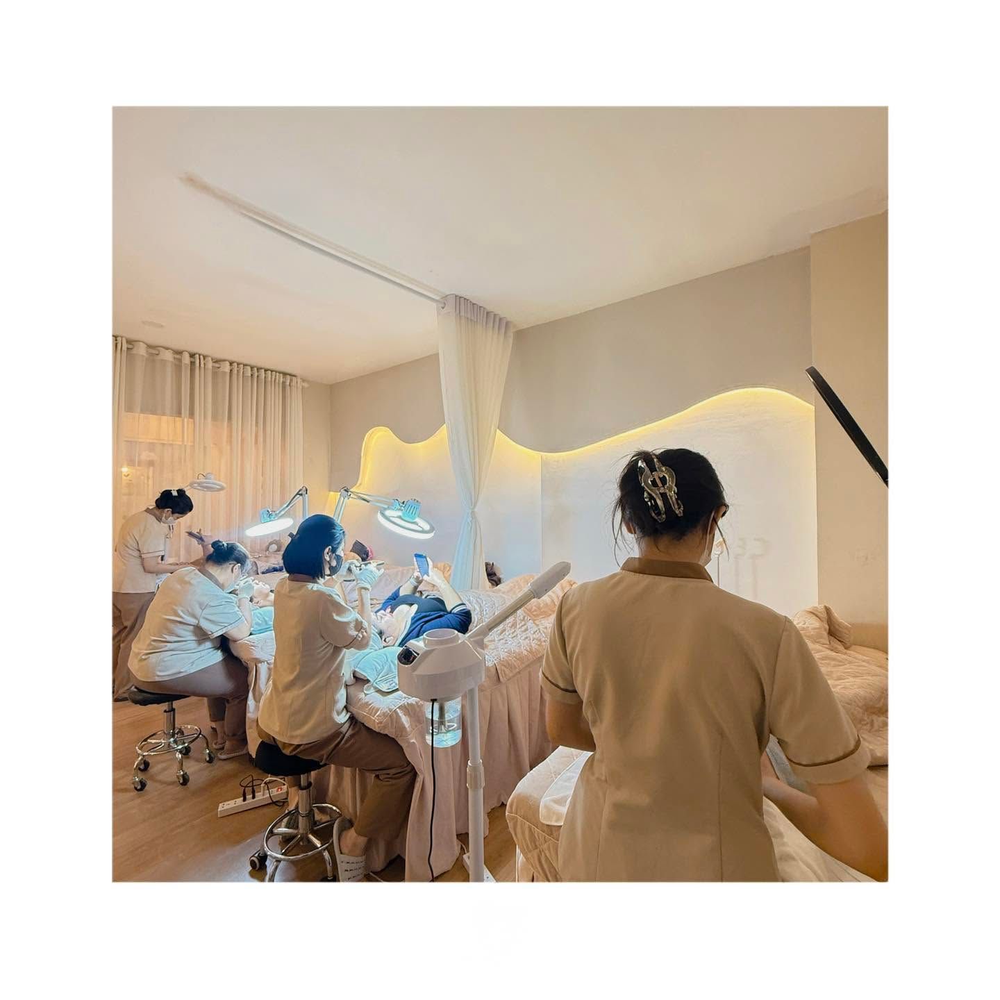 Kỹ thuật viên làm dịch vụ cho khách nằm trong phòng spa ánh sáng ấm, có đèn lúp và xe đẩy dụng cụ