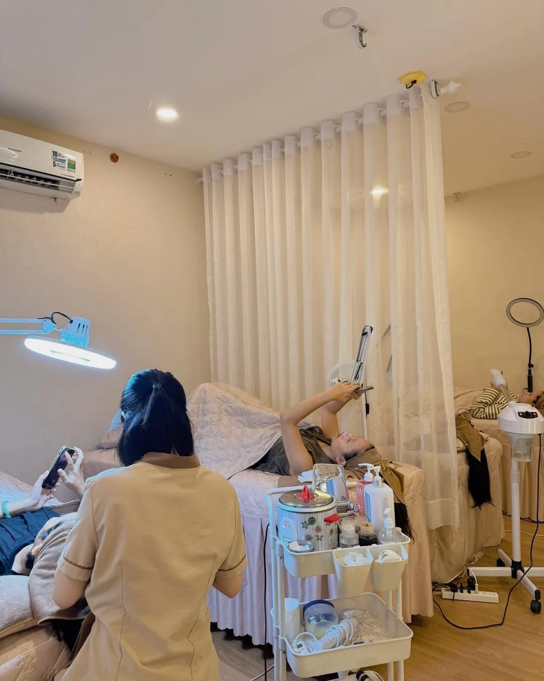 Kỹ thuật viên áo nâu đứng cạnh giường, khách nằm thư giãn trong phòng rèm voan trắng