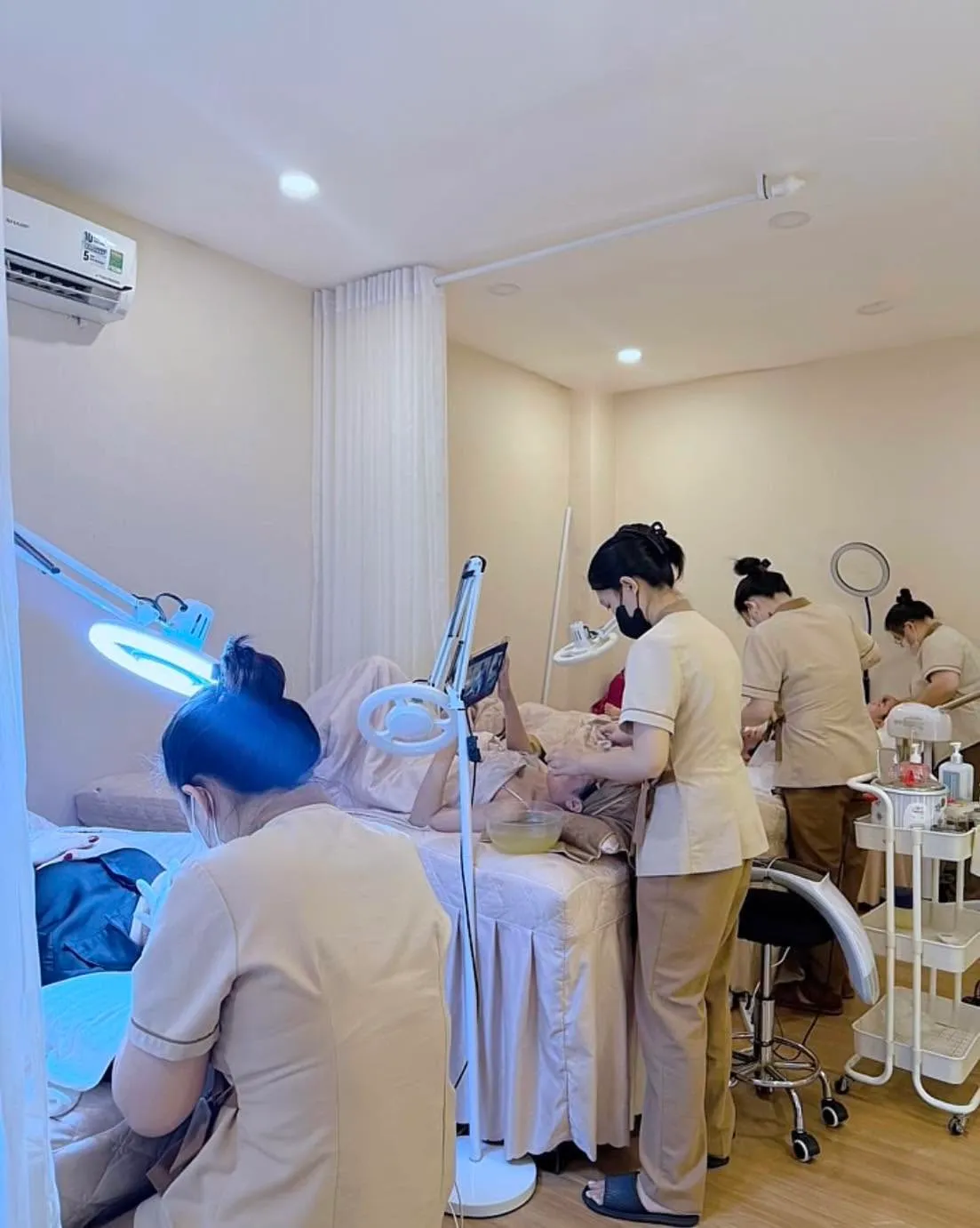 Kỹ thuật viên áo be làm dịch vụ cho khách nằm dưới đèn soi trong phòng nhiều giường